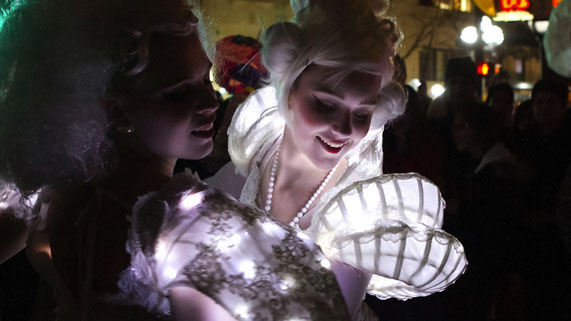 Two people in illuminated, elaborate costumes with white wigs at a nighttime event.