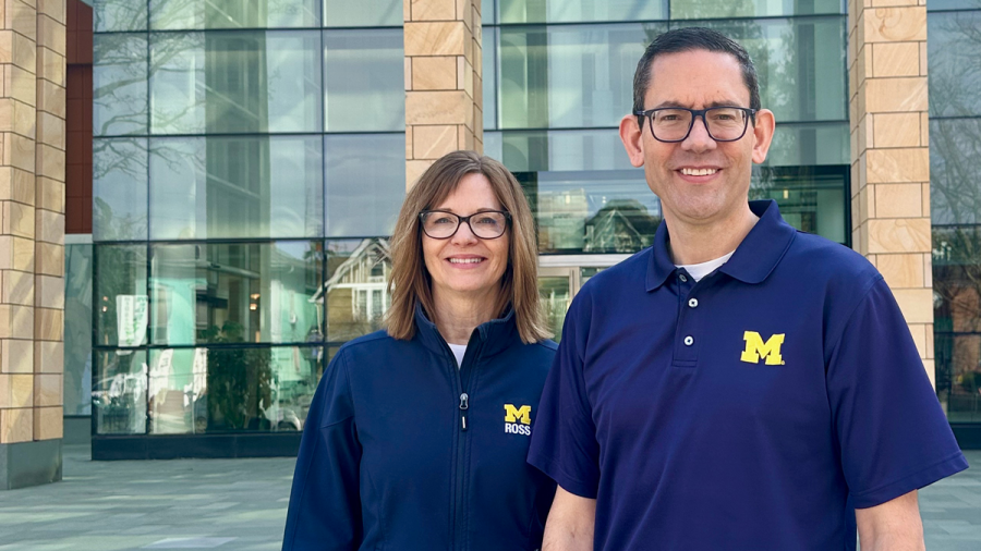 Two people wearing University of Michigan shirts stand smiling in front of a modern building.