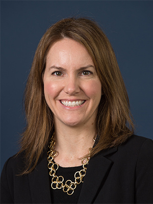Smiling woman in a black blazer with a gold necklace against a dark blue background.