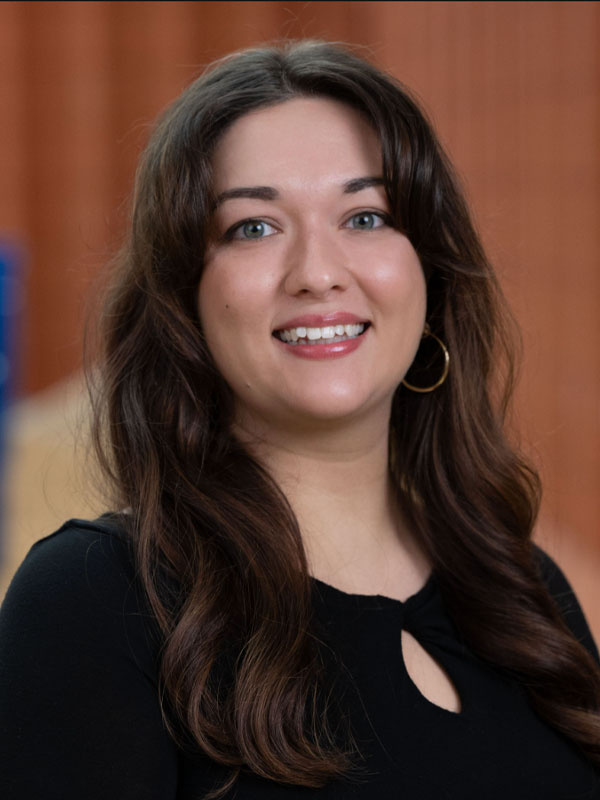 Professional headshot of a smiling woman with long dark wavy hair wearing a dark top.