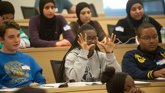 Students seated in a classroom, with one gesturing expressively while speaking.