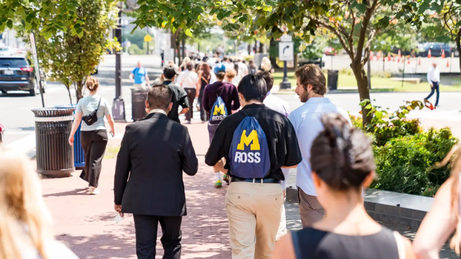 Students walk along a tree-lined campus pathway, one wearing a University of Michigan jacket.