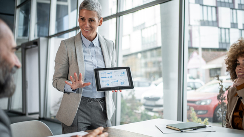 Person in a beige blazer presents a tablet to colleagues in a bright office meeting room.