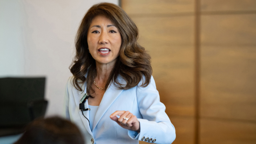A woman in a light blue blazer gestures while speaking in a meeting.