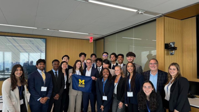 Group of students from the University of Michigan Ross School of Business holding a Michigan Ross maize and blue flag standing in a building in New York City. 