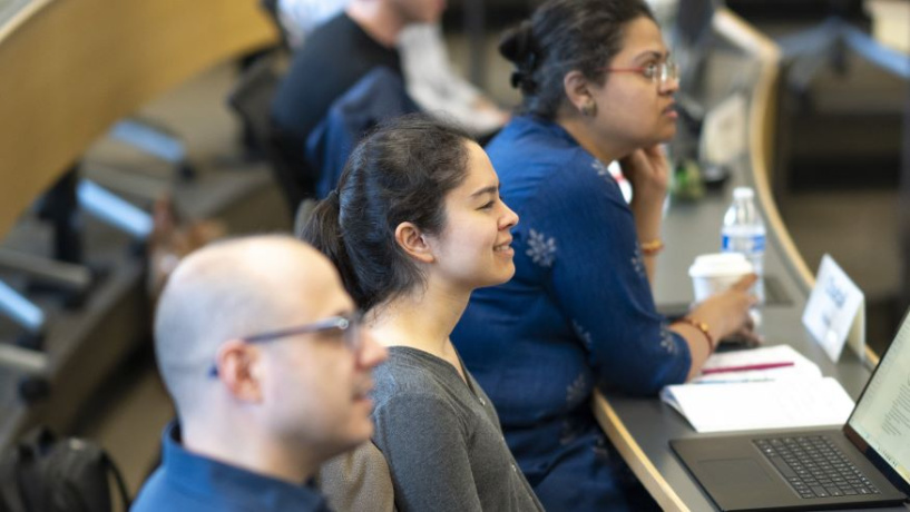 Photo of three Michigan Ross Weekend MBA students sitting in a classroom. There are two women and one man sitting at their desks with laptop computers in front of them. 
