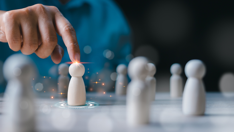 A person’s hand points toward a single wooden figure on a table, highlighting it with a glowing light and digital spark effects. The illuminated figure stands out among several similar, unlit wooden figures in the background, symbolizing leadership, selection, or recognition.