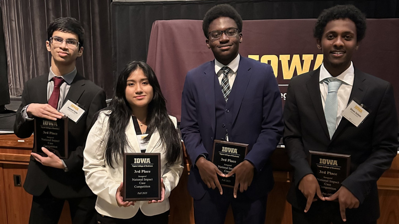 Photo of the four Michigan Ross BBA students who participated in the National Impact Case Competition. There are three male students and one female student. They are holding their third place plaques. Two of them are making the Block M sign with their fingers in the shape of an M. They are all looking at the camera and smiling. The brown and yellow Iowa University banner is in the background. 