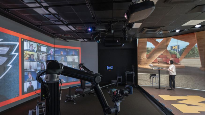 Image of the Michigan Ross digital education studio. There is a large video camera pointing at a professor standing in a large open space and there are multiple students images displayed on a large screen. 