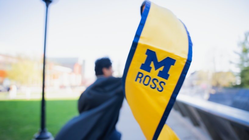 Michigan Ross graduate holds a maize and blue Michigan Ross graduation scarf. The graduate is blurry in the background and the scarf is in focus in the foreground.
