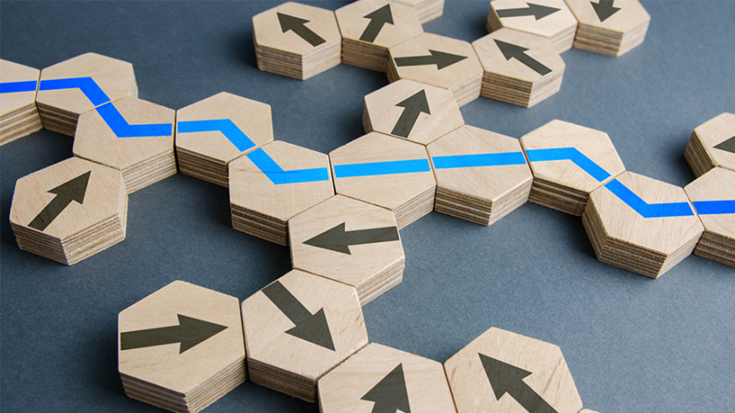 Wooden hexagonal tiles arranged like a network, each printed with directional arrows, with a blue line tracing a clear path through the intersecting routes, symbolizing strategy and decision-making.