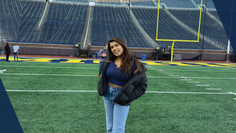 Person smiling and standing on a football field with stadium seats in the background.