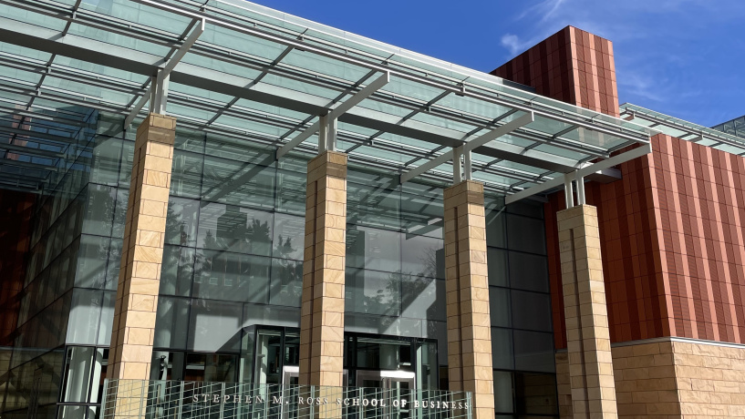 Exterior photo of the Ross School of Business building with four pillars, terra cotta paneling, and glass ceiling. The sky behind it is bright blue. 