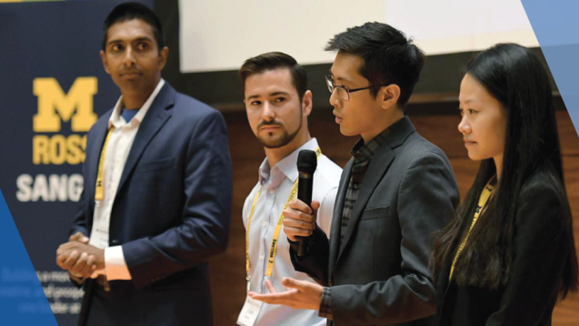 Four people stand together at a University of Michigan Ross School event, with one speaking into a microphone.