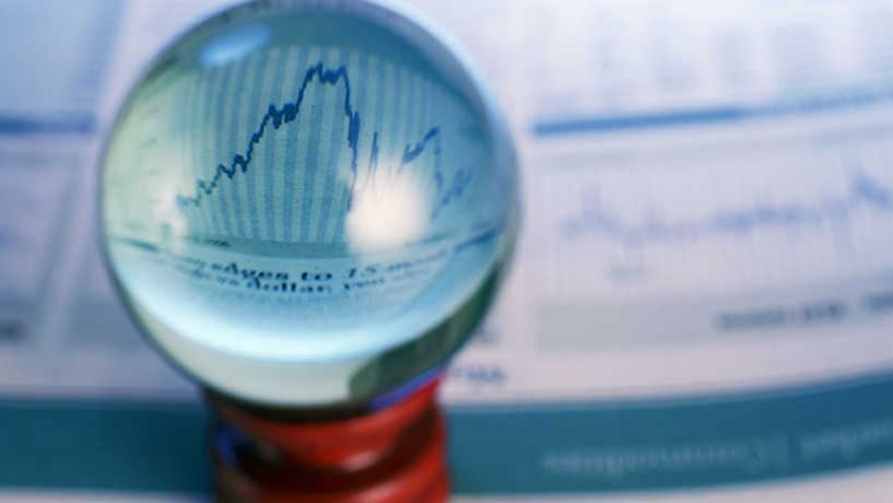 A clear glass sphere sits in front of financial charts, enlarging a section of a blue line graph. The background is intentionally blurred, emphasizing the magnified market trend and suggesting analysis or economic uncertainty.