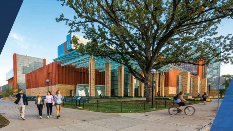 People walking on a pathway outside a modern building with large windows and orange accents, surrounded by trees.