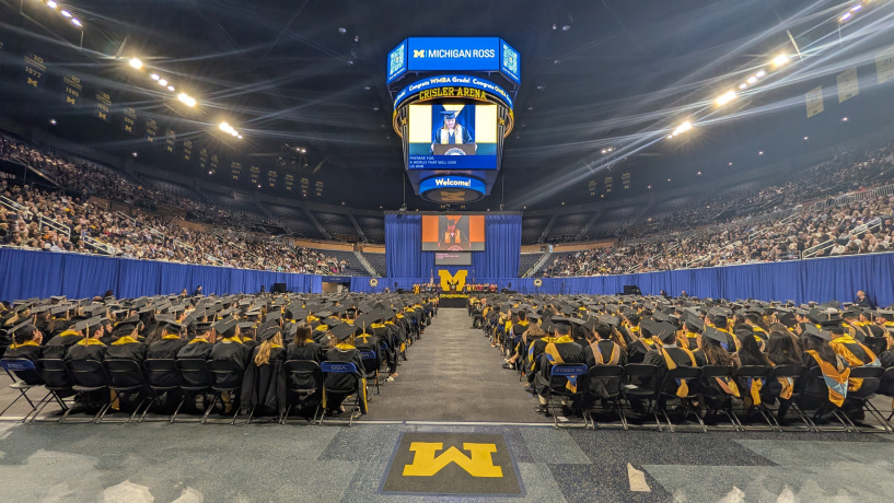 Photo of students in Michigan Ross graduation regalia seated at the Crisler Center with backs to the camera