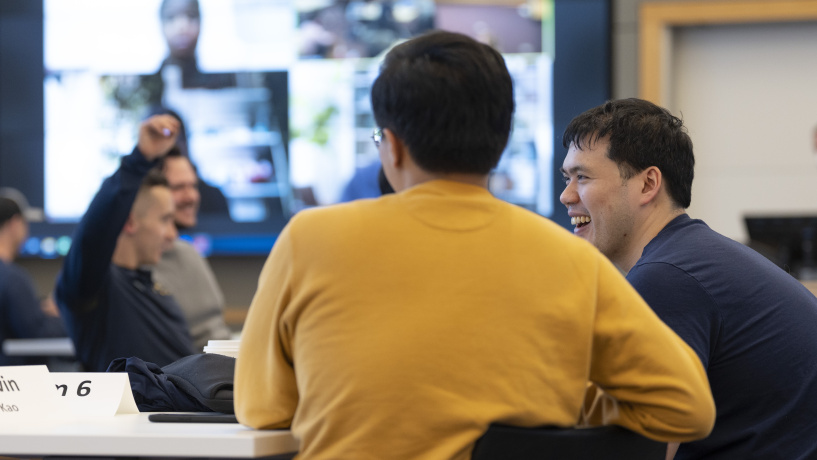 Michigan Ross students in a classroom. One student has his back to the camera and is wearing a maize sweatshirt. Sitting next to a guy in a blue sweatshirt who is smiling. There is a student in the background with his hand raised. There is also a computer screen in the background where some students are online. 
