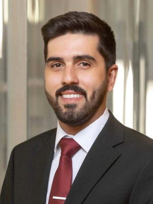 Professional headshot of a man with short dark hair and a beard, wearing a black suit, white shirt, and red tie, smiling in an indoor setting with glass walls in the background.