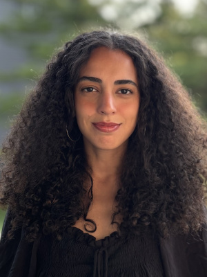 Aline is a black light-skinned woman with curly hair. She is facing the camera and wearing a black blouse.
