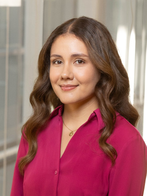 This is a professional headshot of a young woman with wavy brown hair, wearing a fuchsia blouse and a gold necklace. She has a soft smile and is posed in front of a blurred indoor background with glass and light.