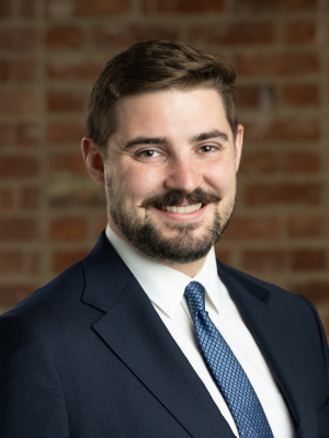 A professional headshot of man in a navy suit jacket facing right and smiling at the camera. He is white with dark brown hair, brown eyes, and a mustache and beard. He wears a white shirt and a blue tie.