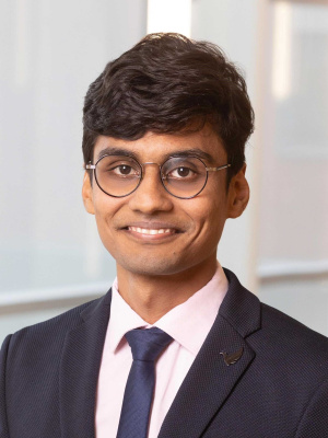 Professional headshot of Amogh Kabra in a suit and tie, wearing glasses and smiling, in a corporate office setting