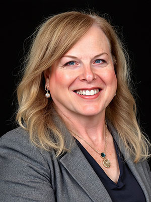 Chrissie Watchter with long blond hair wearing a gray blazer and dark shirt against a black background.