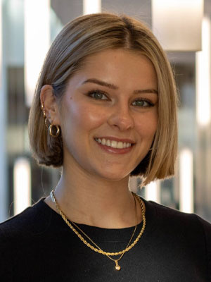 Headshot of Emma Sharpe: A person with short blond hair smiling indoors with blurred lights in the background.