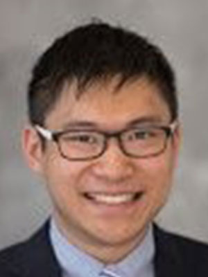 Bennet Lai wearing glasses and a suit jacket against a gray background.