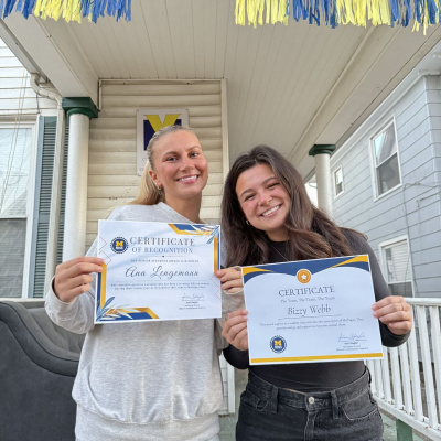 Two women pose smiling with certificates on the porch of a house with a maize and blue banner.