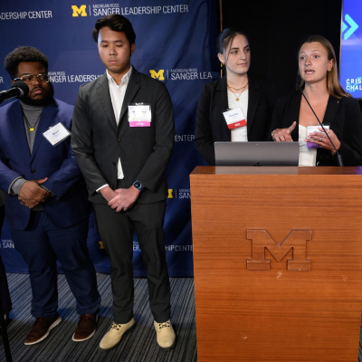 Four students in business attire present at a podium with the University of Michigan logo.