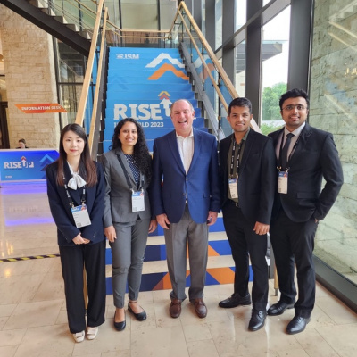 Five people in business attire stand in front of a staircase with a "RISE" conference banner.