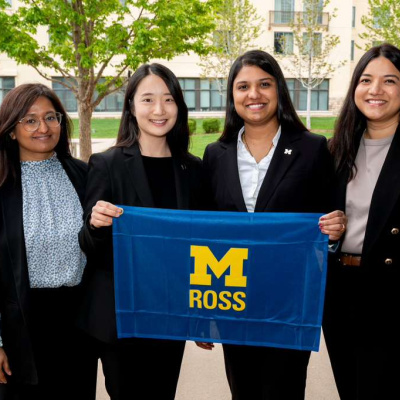 Four women in business attire hold a University of Michigan Ross banner outdoors.