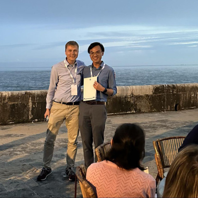 Two people stand by the ocean at sunset, one holding a certificate during an award presentation.