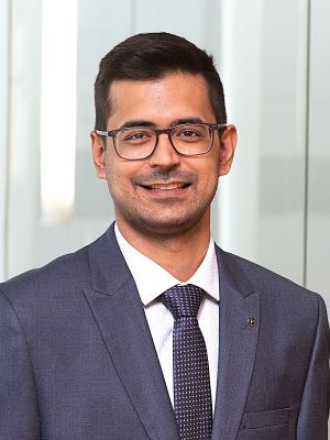 A man with short brown hair wearing a navy suit smiling in a bright room