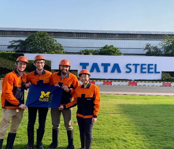 Students in orange hard hats stand in green grass in front of Tata Steel sign and headquarters