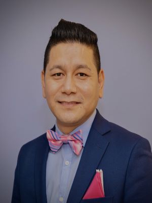 A man with short brown hair smiling in a blue suit jacket and light blue shirt with a pink striped tie