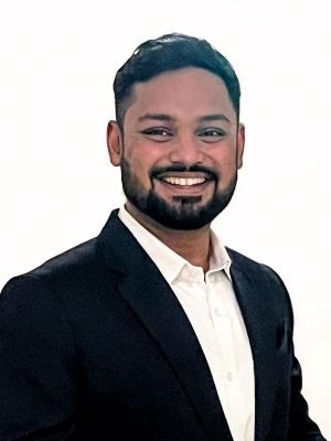 A man with short black hair smiles, wearing a black suit and white shirt in front of a white background