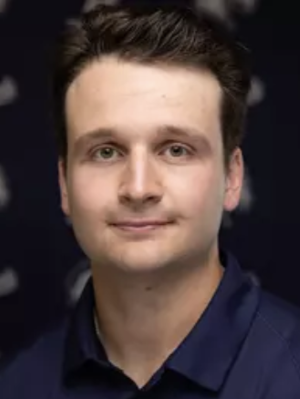 A man with short brown hair smiles for a headshot, wearing a navy blue collared shirt in front of a black background