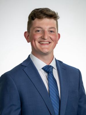 A man with short brown/blonde hair smiles wearing a blue suit, white shirt, and blue tie in front of a white background