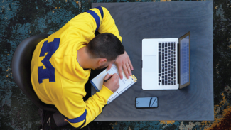 Man sitting at desk writing in a notebook with a laptop nearby.