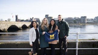 Three female students and one male student standing in Ireland holding a blue and yellow Michigan Ross flag.