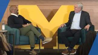 Marcus Collins and Dave Deno sitting in chairs on a stage talking with a large block M in the background