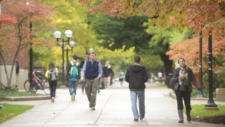 Photo of the University of Michigan campus with male and female students walking in the fall season. The trees have leaves that are green and orange.