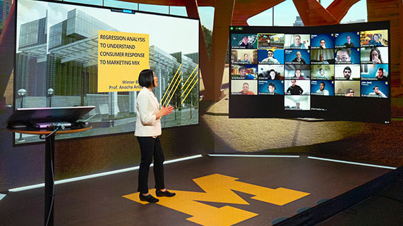 professor standing on digital stage with block "M" in front of Zoom class giving presentation